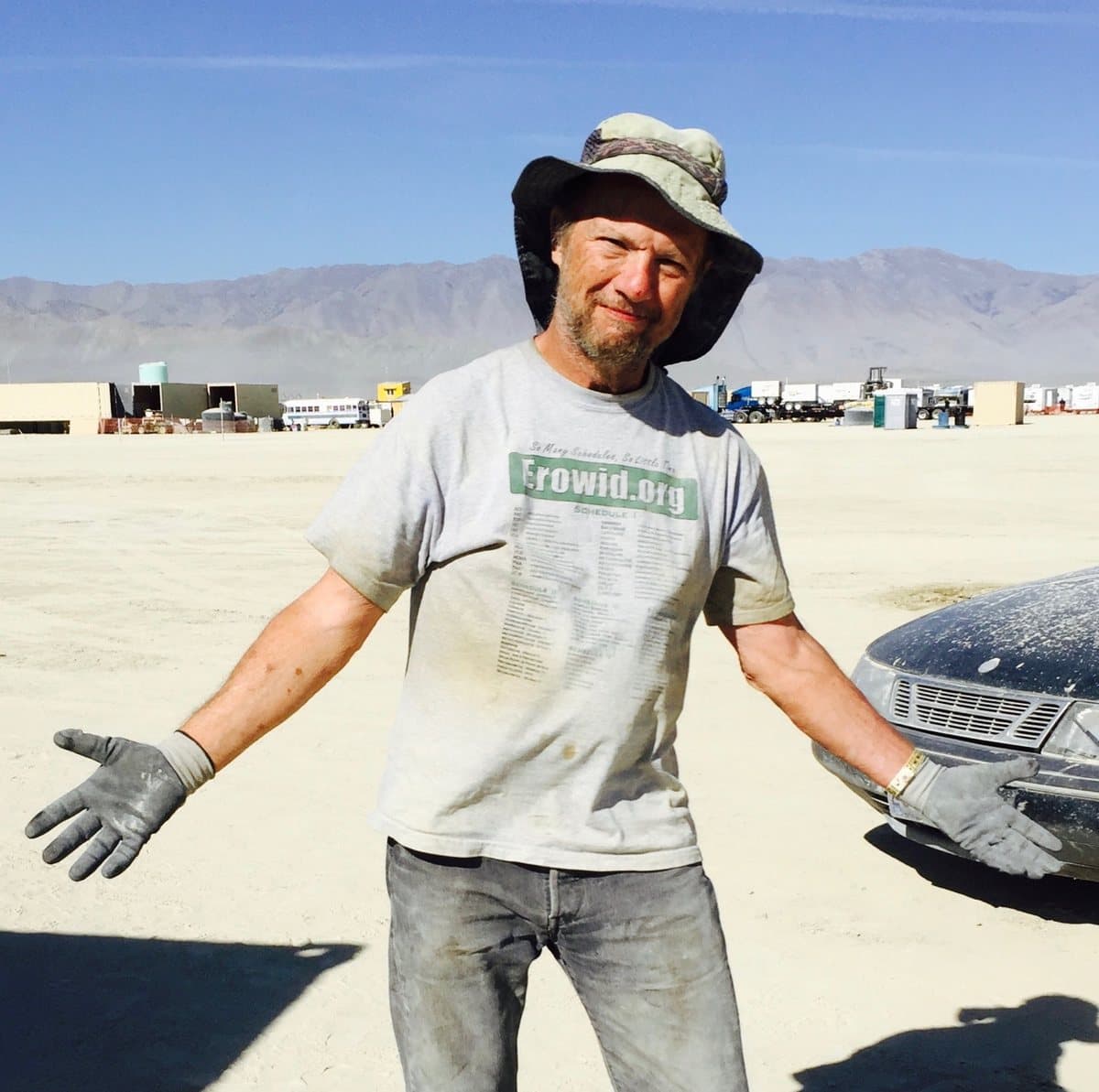 Joe Weisman on the playa in a wide-brimmed hat and dusty work gloves, arms open in a shrug, a dust-covered car beside him.